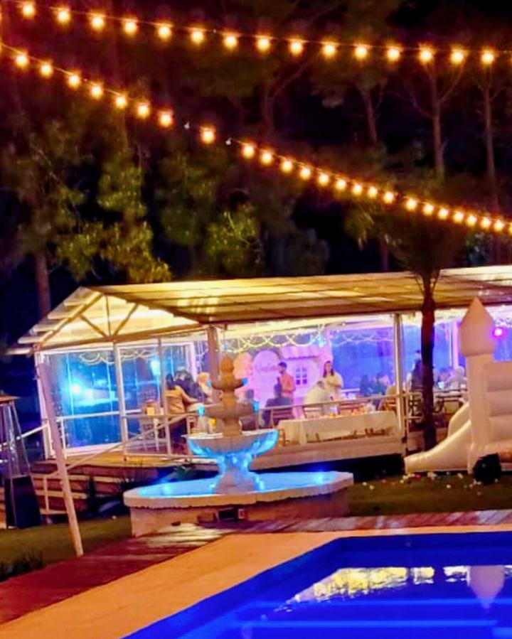 Salón de eventos vidriado de noche, con guirnaldas de luces y fuente